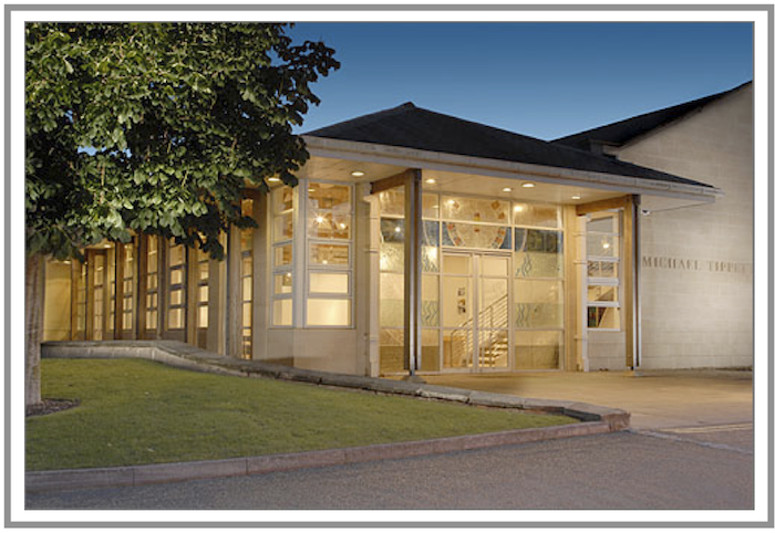 Frontage of a glass-walled building with one storey and angular roof attractively pictured at dusk with lights shining from the glass fronted interior.